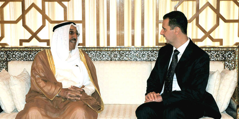 Two men, one in Arab Gulf dress and another in a black suit with tie, sit on a couch in conversation