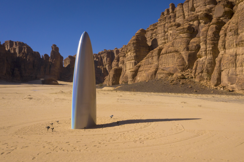 large silver bullet shaped art installation protruding vertically from ground with brown sand and cliffs in the background