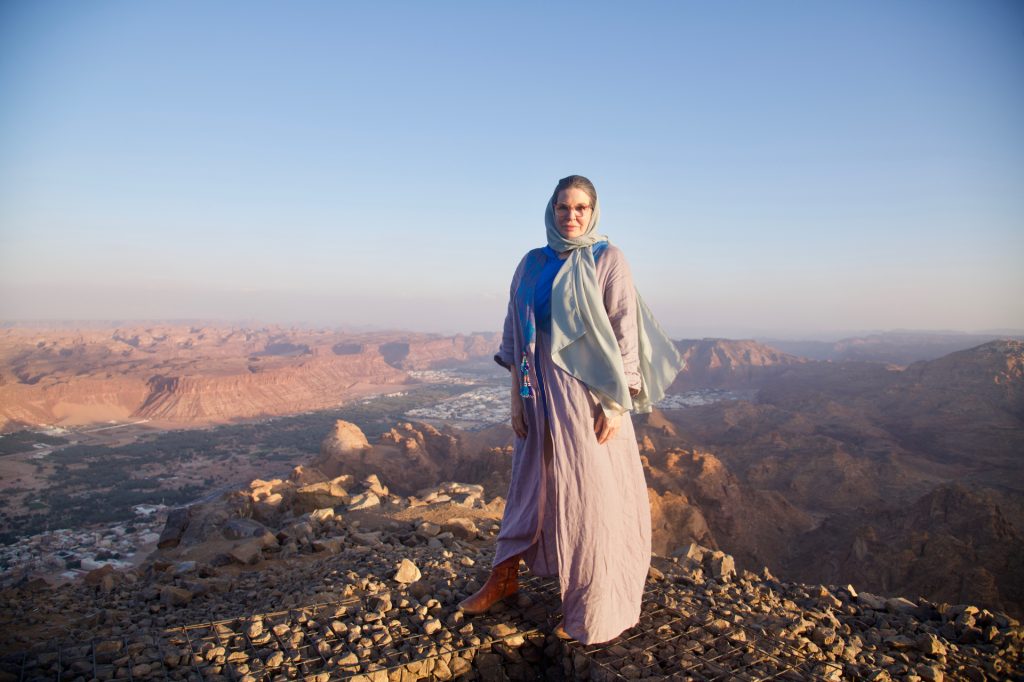 Woman wearing a blue abaya standing at a cliff edge with buildings in the valley below