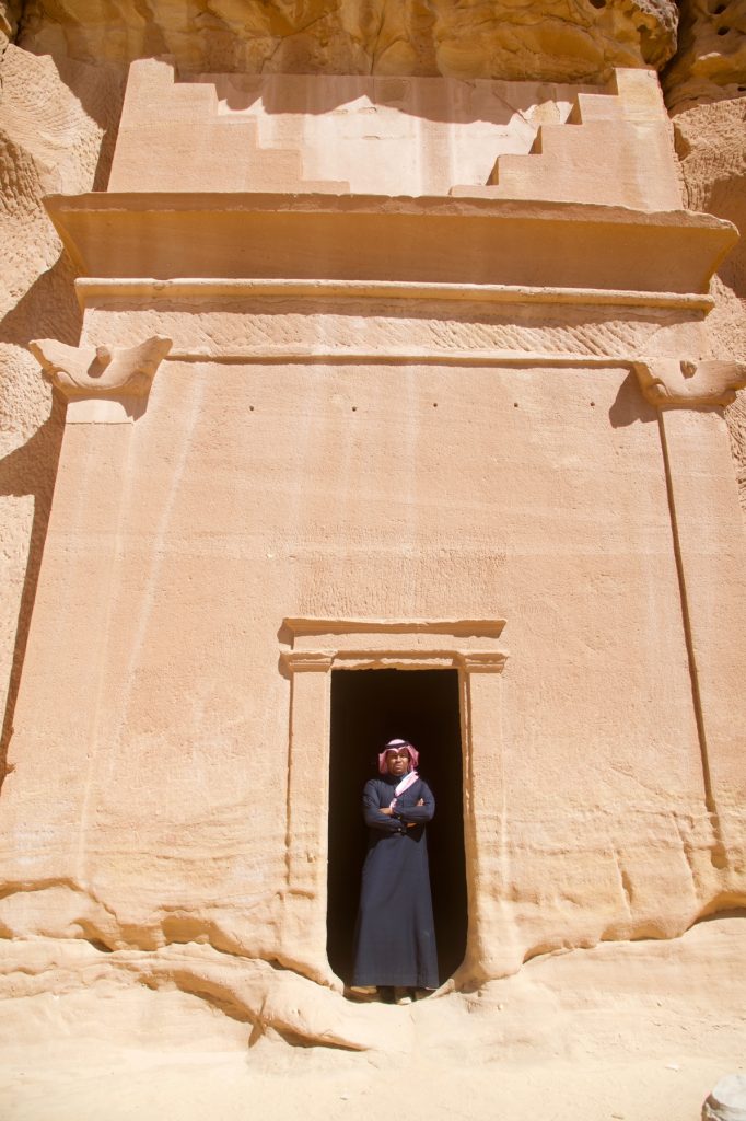 tomb carved into a rock cliff with a man wearing a black bisht standing in the doorway