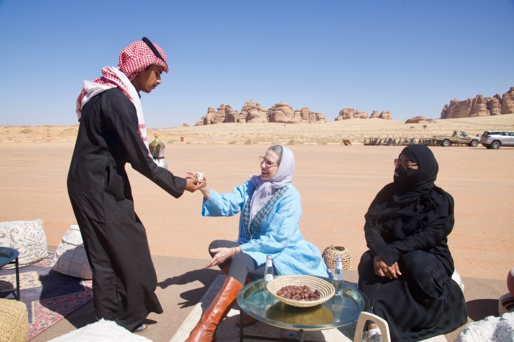 a woman in a blue abaya is seated next to a woman in a black abaya and niqab and served a drink by a man in a black thobe
