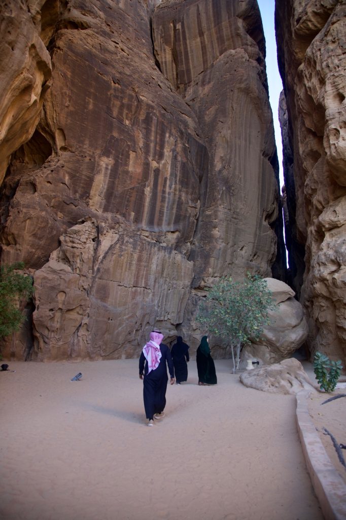 Several individuals wearing black thobes and abayas walk toward a narrow opening in a rock cliff.