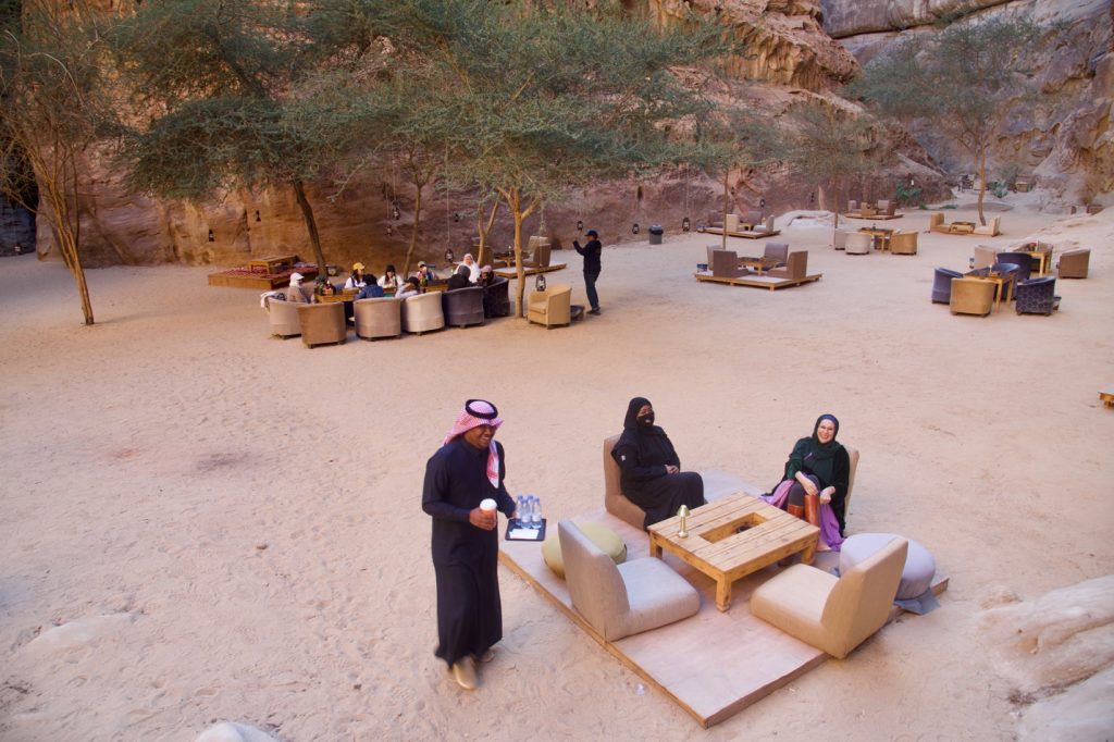 several restaurant tables, some occupied, sit on a sandy desert floor with rock cliffs as walls