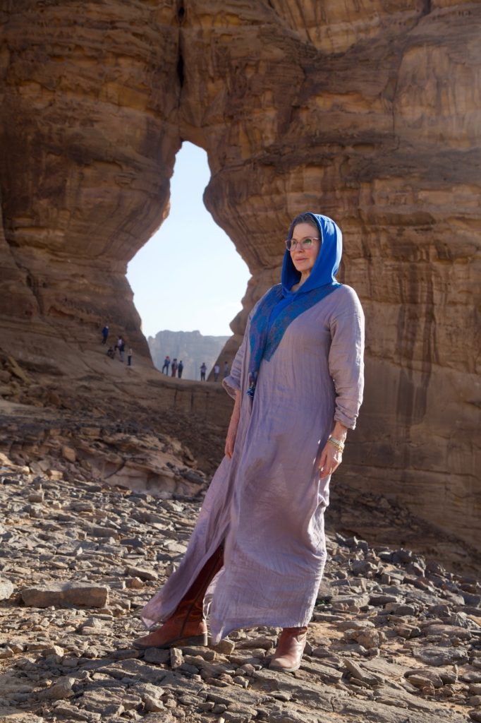 a woman in a blue abaya stands in front of a jar-shaped hole in a rocky cliff