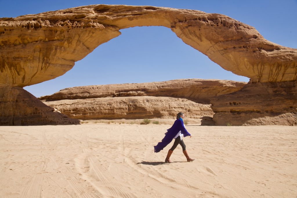 a large rock arch in the desert with a woman in a blue abaya walking in front of it