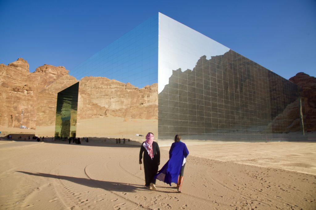 a man in a black thobe and a woman in a blue abaya walk toward a large mirrored building in a desert adjacent to rocky cliffs