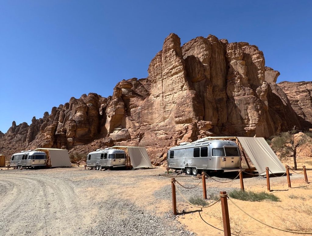 shiny gray airstream trailers sit on a gravel road beneath rocky cliffs and a blue sky