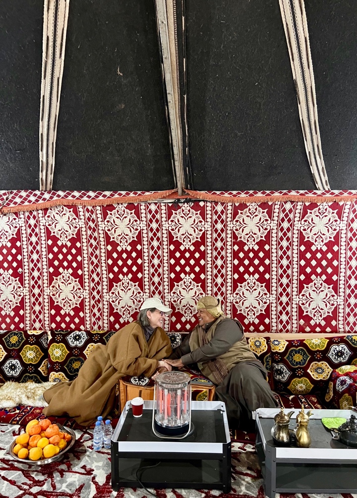 a man and a woman sit in conversation while smiling inside a large tent with red and white upholstered walls