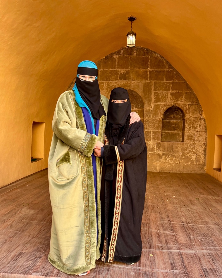 two women, one in a black abaya and the other in a blue and green abaya, both wearing a face covering and standing with their arms around each other facing the camera