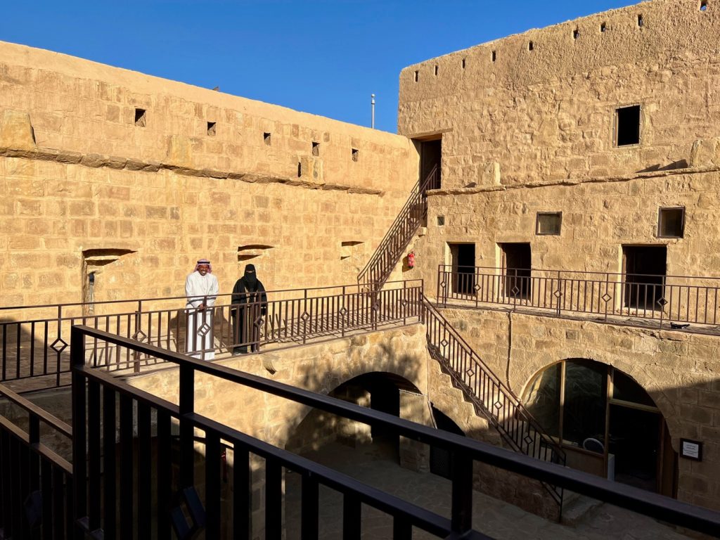 courtyard surrounded by stone walls with two individuals standing on a second floor ledge with stairways leading up and down