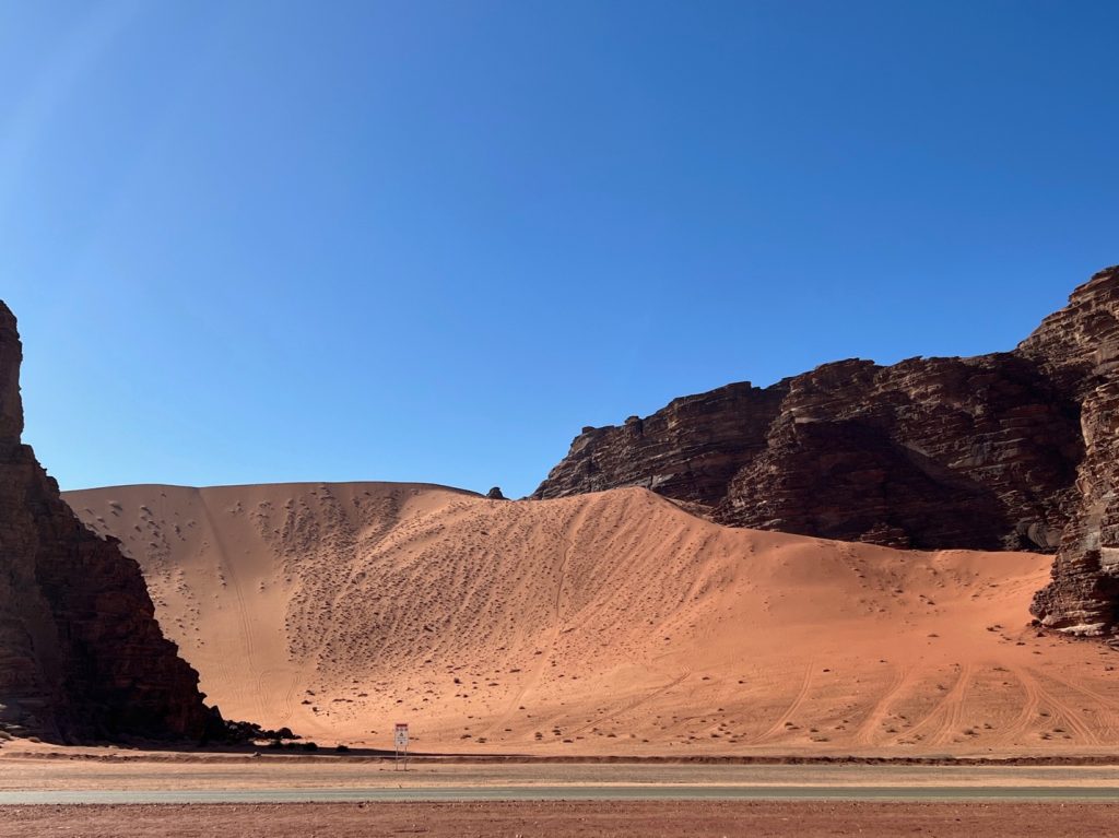 a red sand dune with tire tracks on it and rocky cliffs behind it
