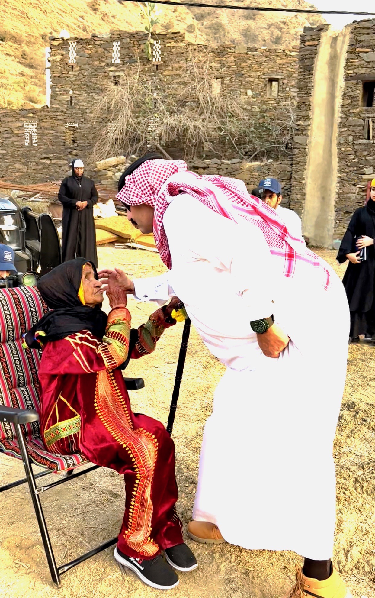 an elderly woman seated in a chair is greeting by a man wearing a white thobe