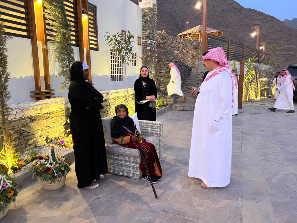 a man in a white thobe speaking to two women in black abayas standing and an elderly woman in a multi-colored dress who is seated