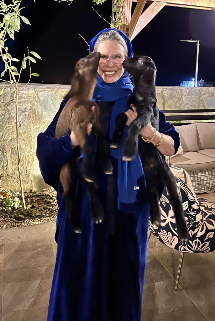 woman in blue abaya smiling while she holds two goat kids