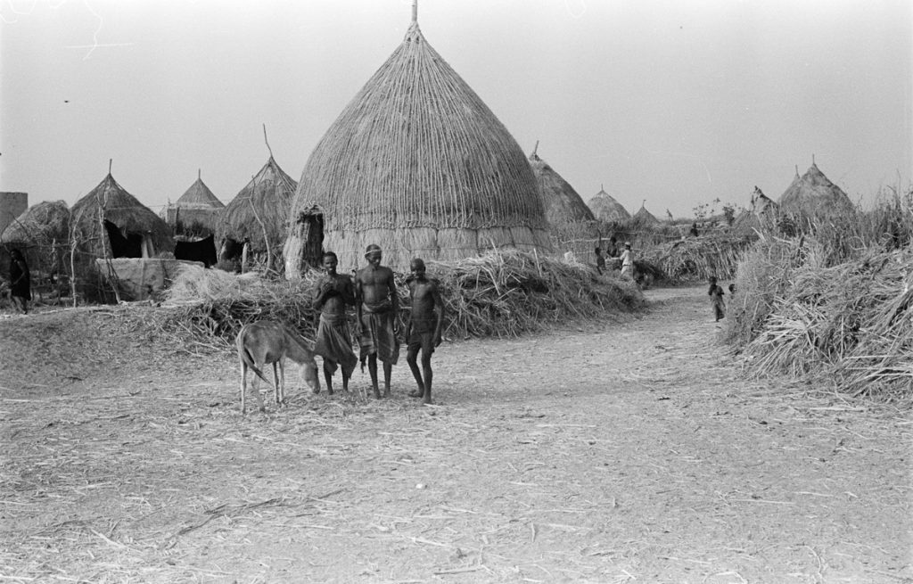 View of round huts with pointed roofs in the town of Umm al Khashab. Three men stand with a donkey in front of the huts.