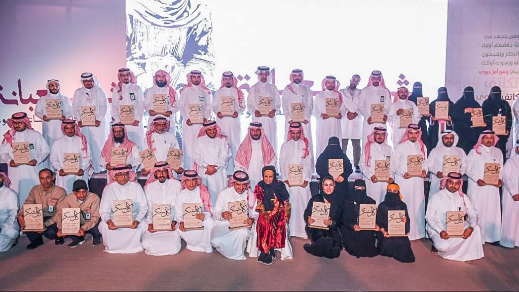 A group of men in white thobes and several women in black abayas gather for a group picture after receiving a plaque