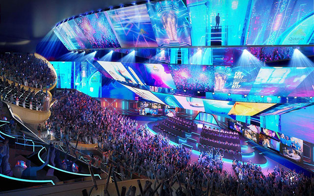 an audience sits in tiered auditorium seating looking down on large video screens above a stage